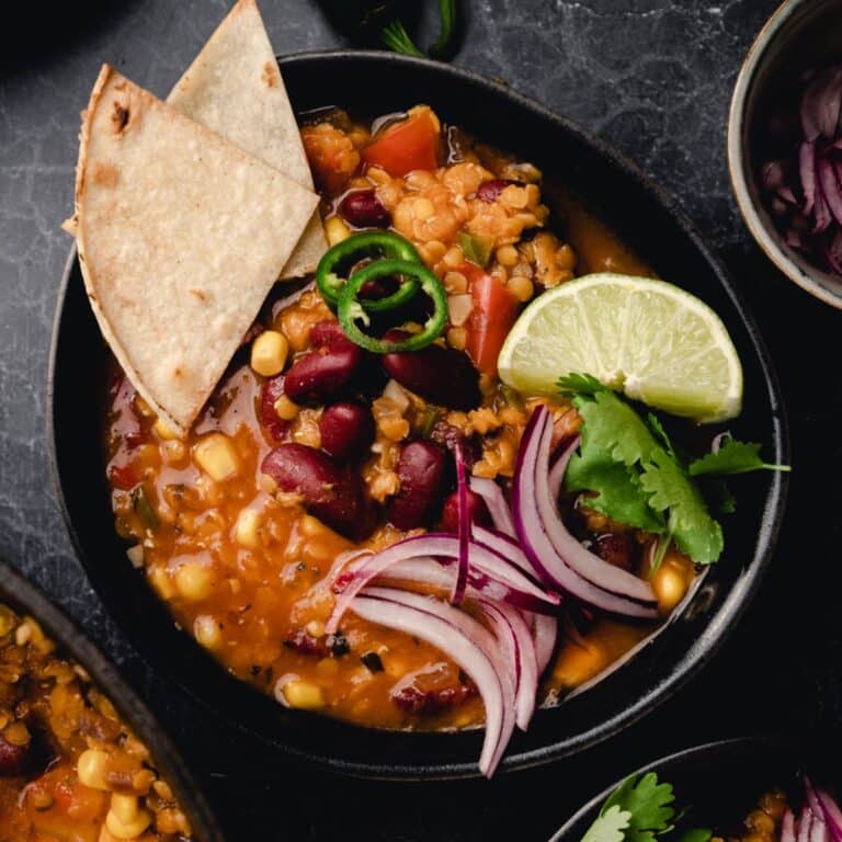 A bowl of vegan chili with corn, kidney beans, red onion slices, jalapeño, cilantro, and a lime wedge. Two tortilla chips are placed on the side.