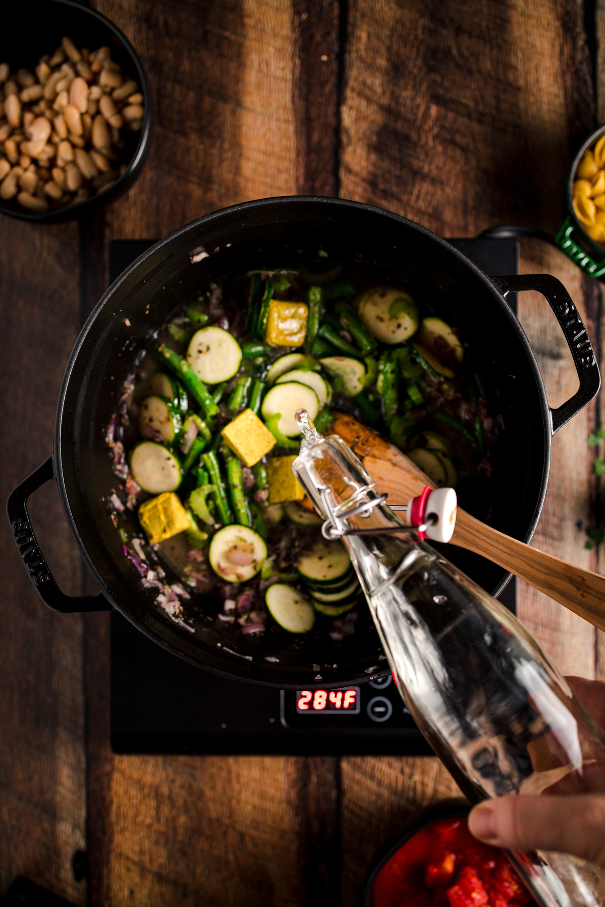 A person stirs vegetables in a pan on a wooden table.