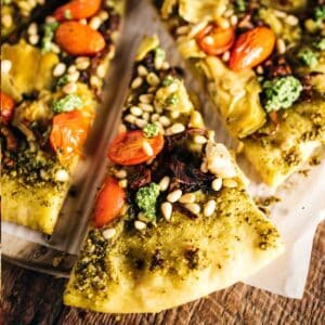 A slice of vegan pesto pizza topped with fresh tomatoes, artichokes, pine nuts, and dollops of green sauce rests enticingly on a wooden surface.