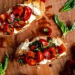 Two pieces of vegan bruschetta topped with cherry tomatoes, fresh basil, and a delightful spread rest on a wooden surface.