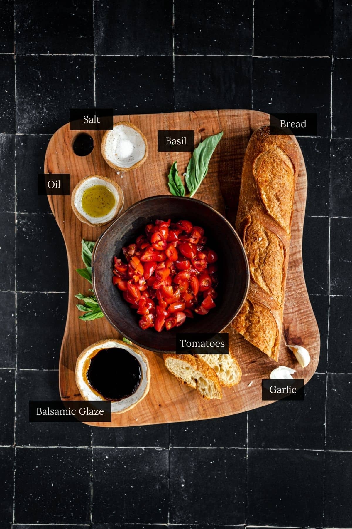 A wooden board showcases vegan bruschetta ingredients: a baguette, a bowl of chopped tomatoes, basil leaves, garlic, salt, oil, and balsamic glaze on a black tiled background.