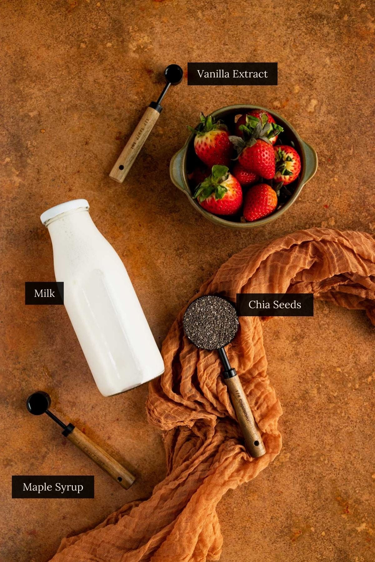 Ingredients on a copper surface include a bowl of strawberries, a bottle of milk, a cup of chia seeds, a measuring spoon with maple syrup, and vanilla extract.