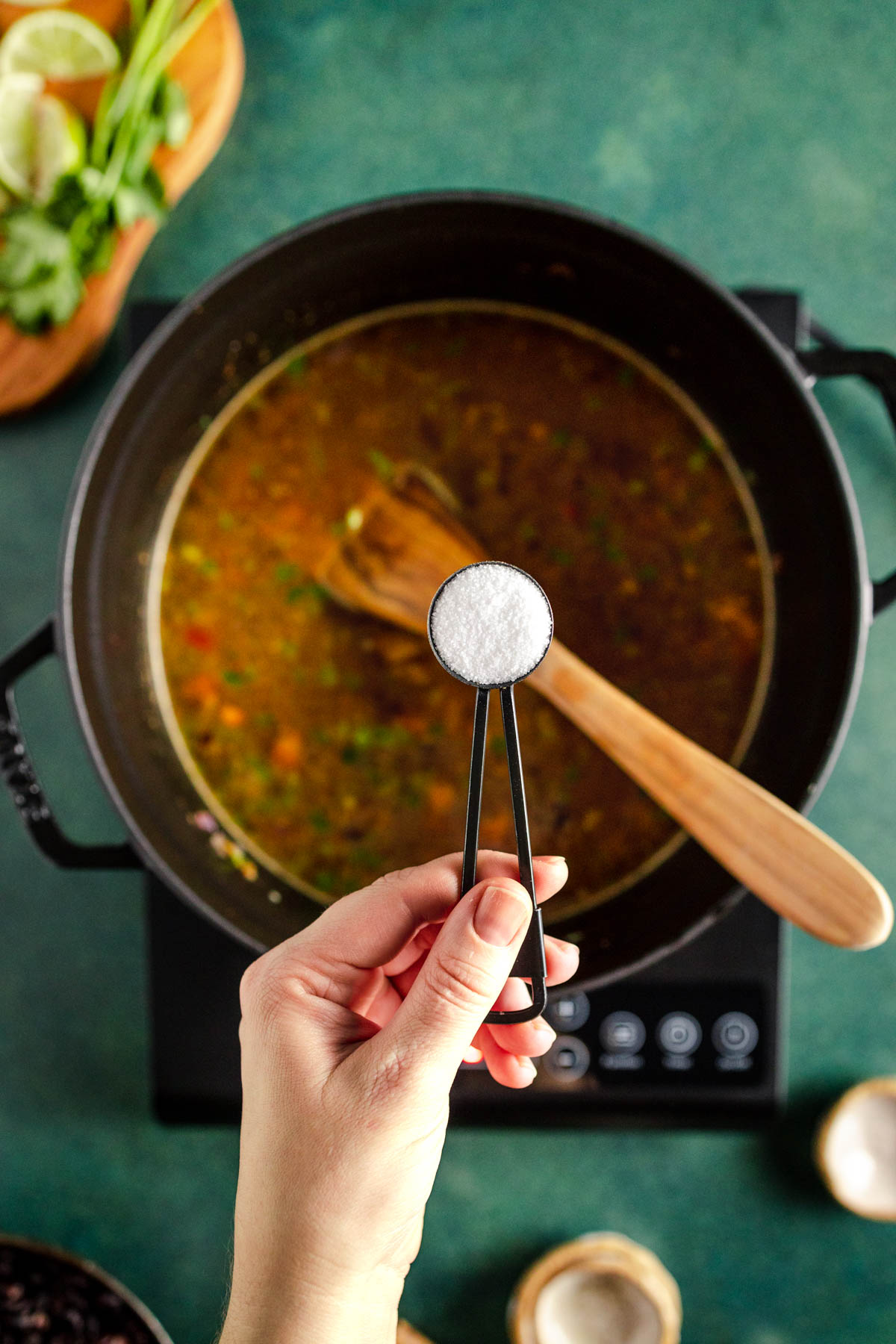 A person holding a spoon of salt over a pot of soup.