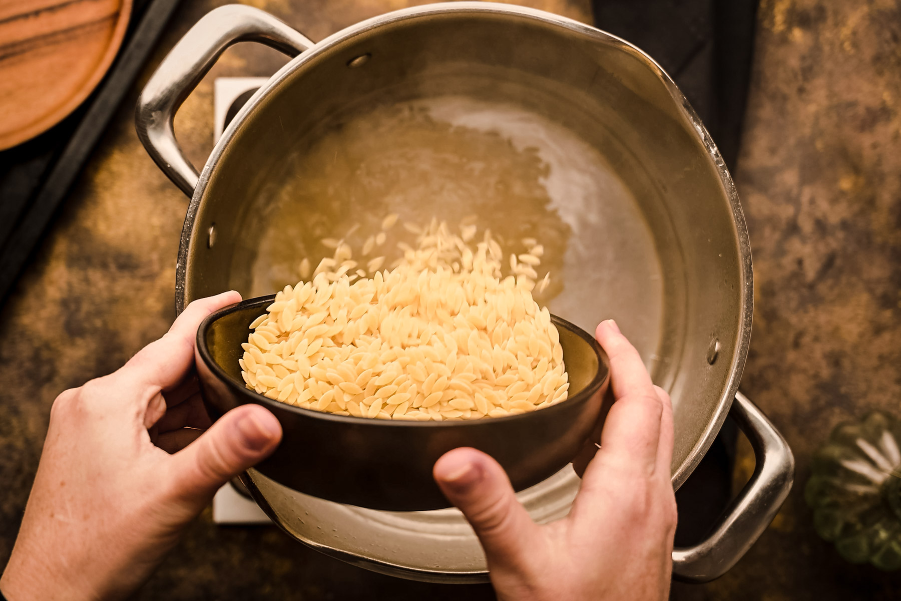A person pouring orzo into a pan on top of a stove.