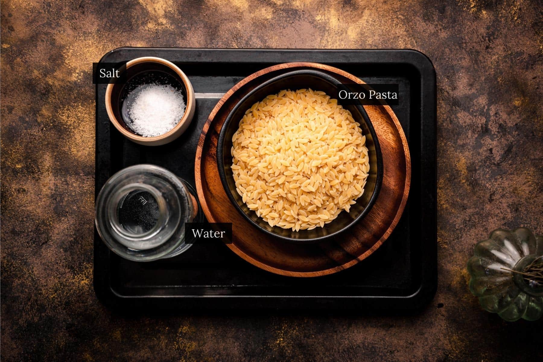 Tray with a bowl of uncooked orzo pasta, a small bowl of salt, and a glass of water on a dark surface.