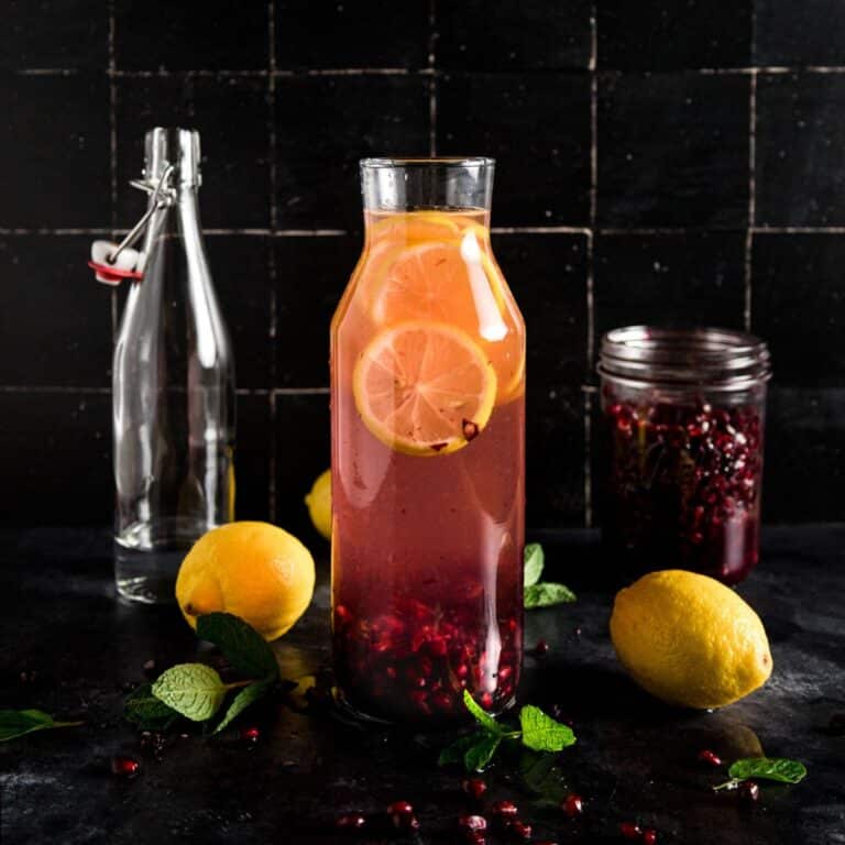 Glass pitcher with pink pomegranate water, lemon slices, and scattered pomegranate seeds, surrounded by whole lemons, fresh mint leaves.