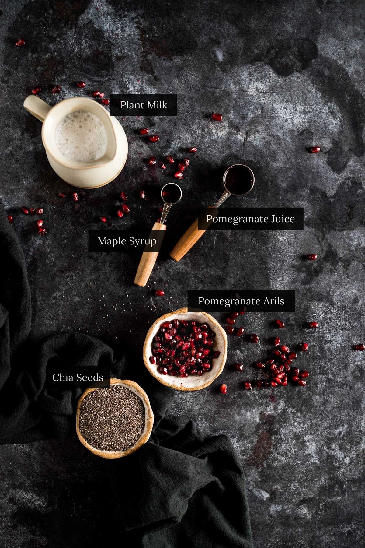 Ingredients for a delicious pomegranate pudding on a dark surface, including plant milk, maple syrup, chia seeds, and pomegranate juice.