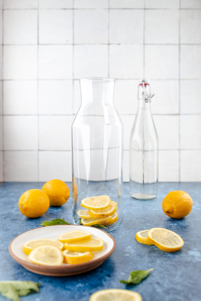 Empty glass pitcher with whole and sliced lemons on a plate, accompanied by a water bottle and mint leaves, on a blue surface with white tiled background.