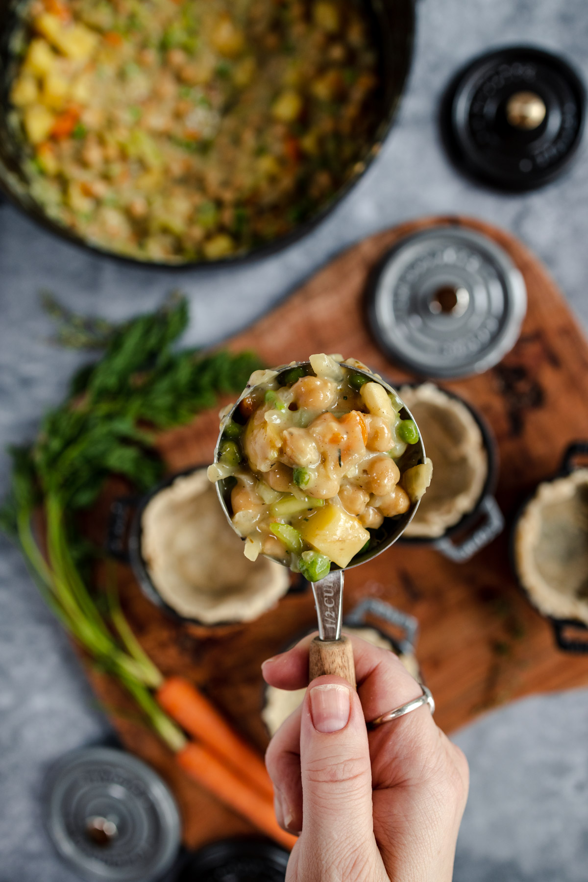A person holding a ladle of chunky vegetable soup over a pot, with ingredients and empty bowls on the table.