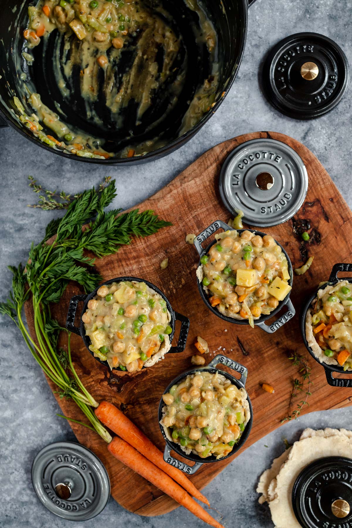 Prepared ingredients for a dish arranged in small cooking pots with fresh carrots and herbs on a wooden cutting board, and an empty mixing bowl with residue.
