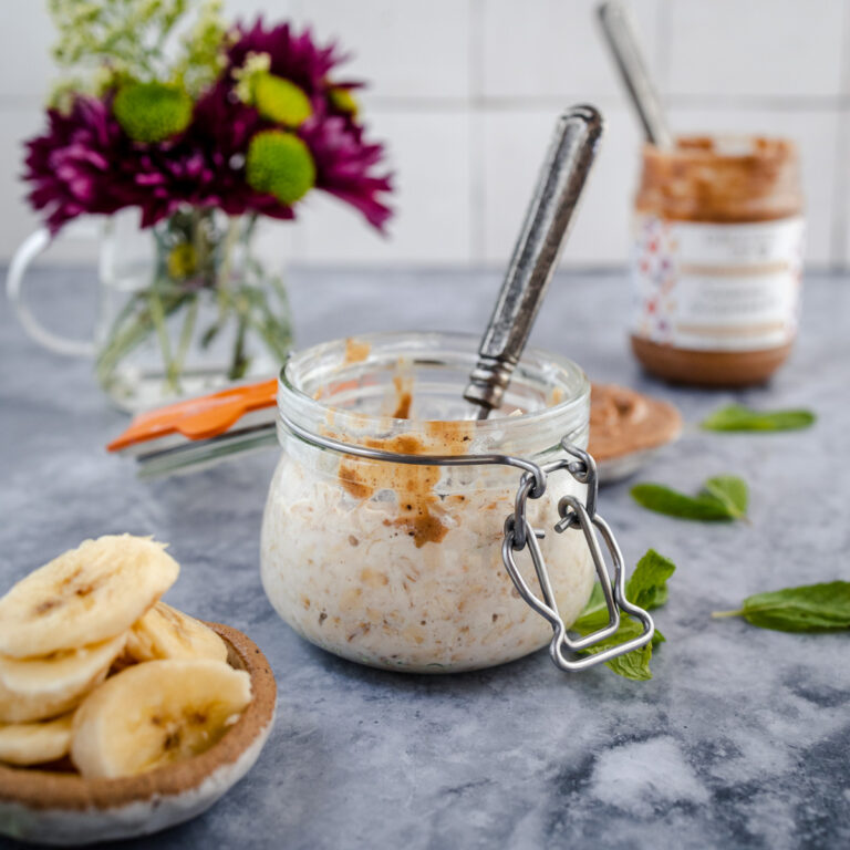 A jar of overnight oats without yogurt, accompanied by sliced bananas and a jar of peanut butter on a marble countertop.