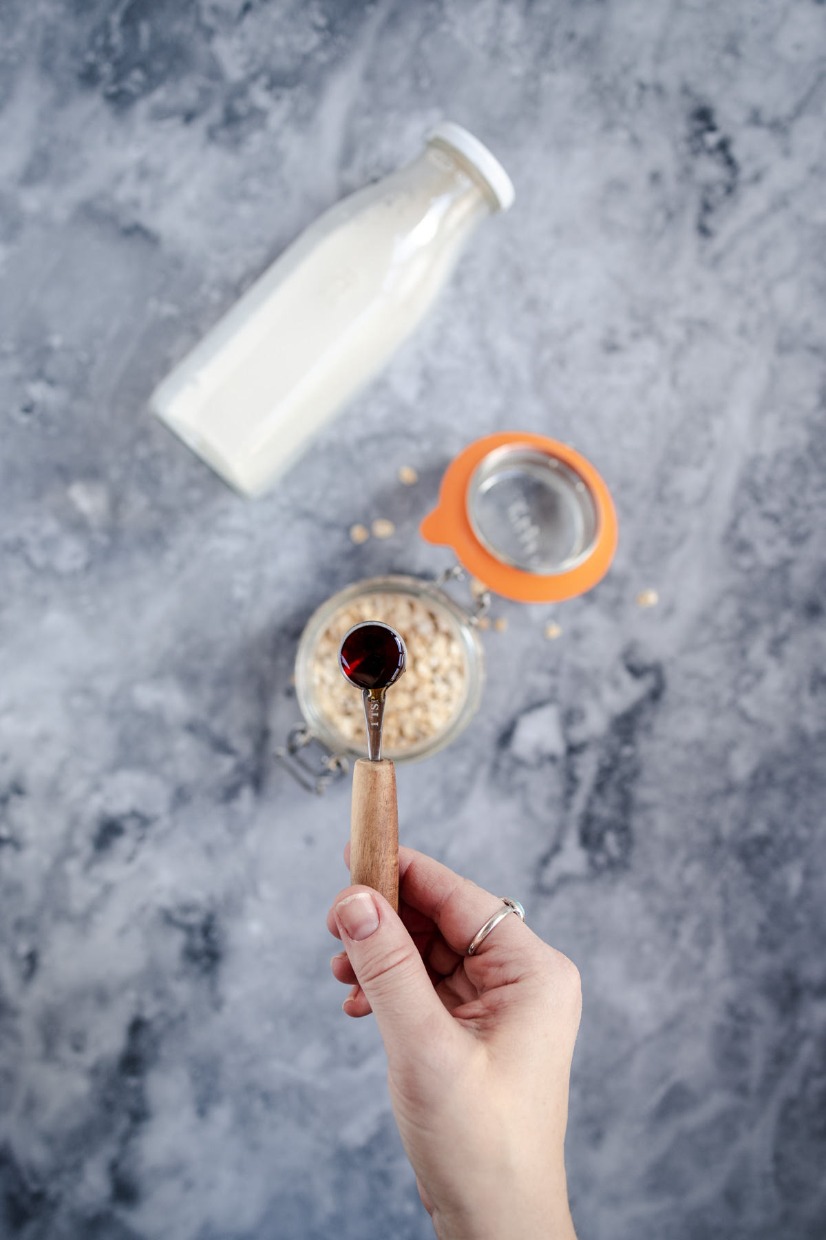 A person pouring syrup into a bowl of oatmeal next to a bottle of milk on a marble surface.