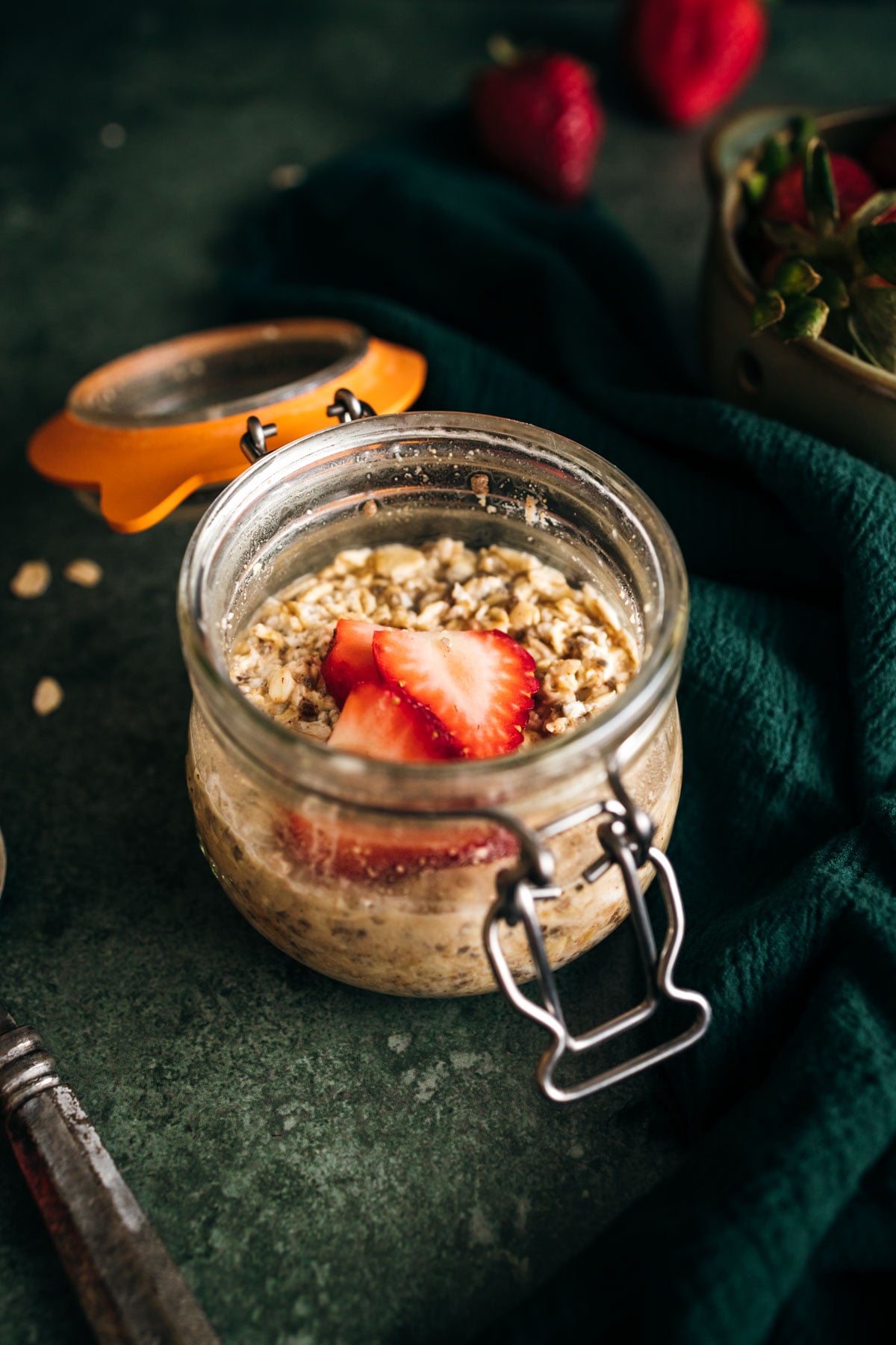 A jar of overnight oats topped with a sliced strawberry on a dark textured surface with fresh strawberries in the background.