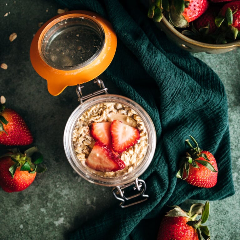 Overhead view of a glass jar filled with oatmeal and fresh strawberries on a dark green cloth, with whole strawberries scattered around.