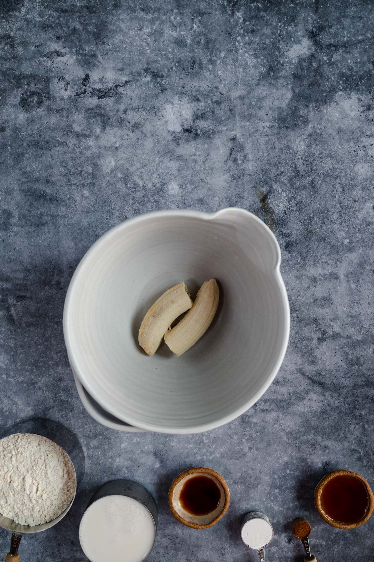 A white bowl with banana slices on a kitchen counter, surrounded by ingredients for baking.