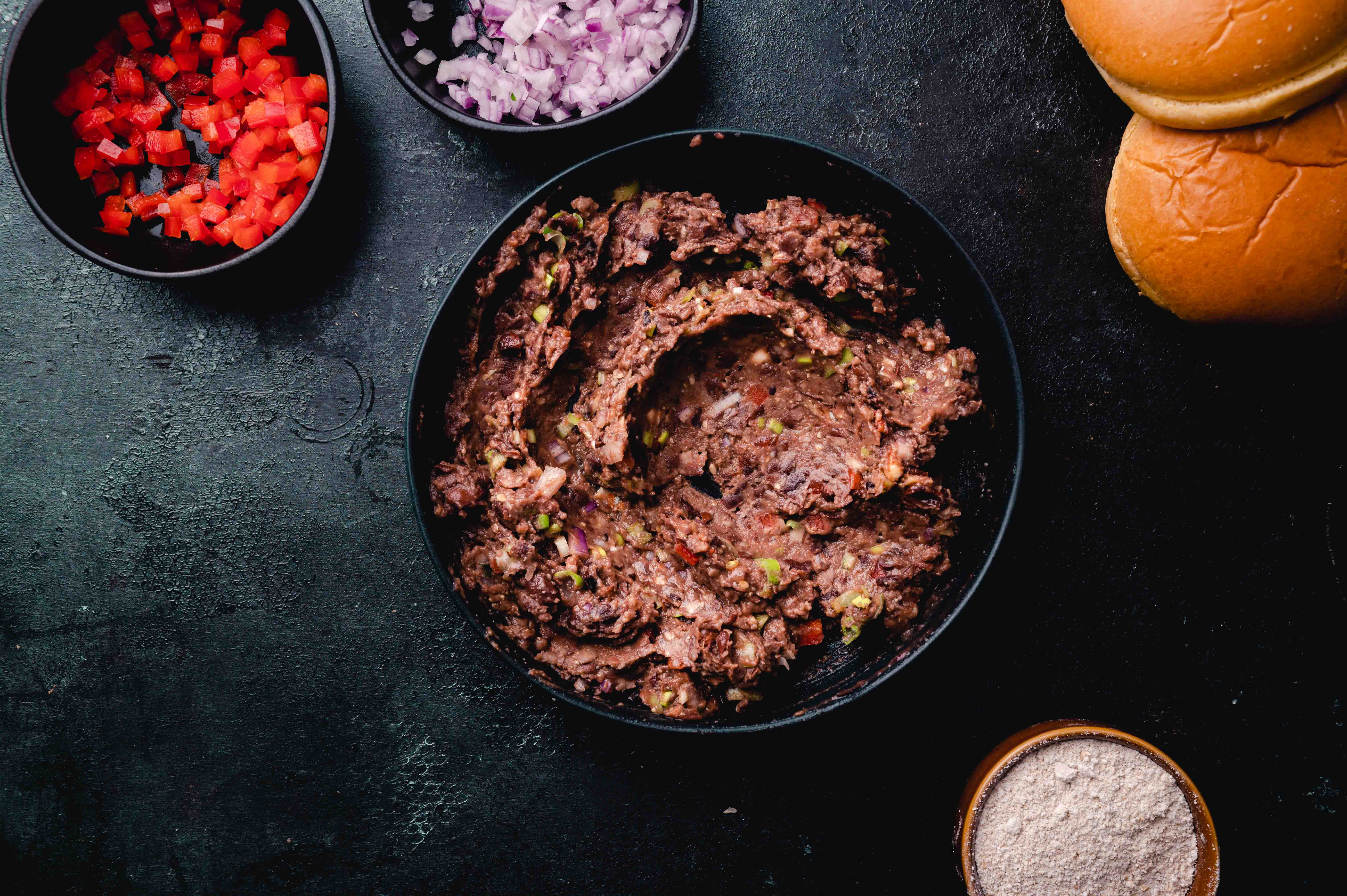 A skillet of cooked ground beef with chopped red peppers and onions, accompanied by buns and seasonings, on a dark surface.