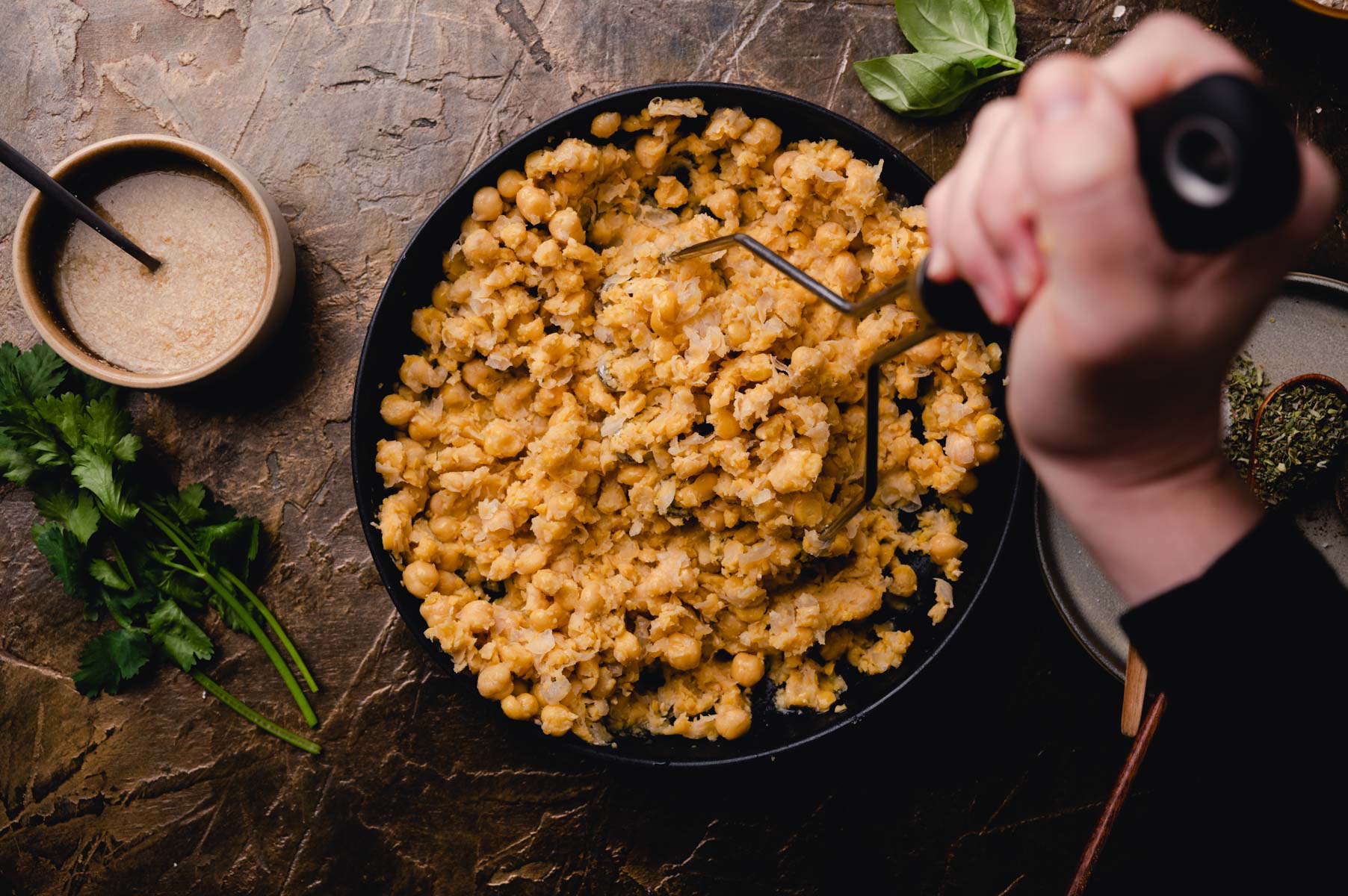 A skillet with sautéed chickpeas garnished with herbs, next to a bowl of sauce and fresh parsley, with a person stirring the dish.