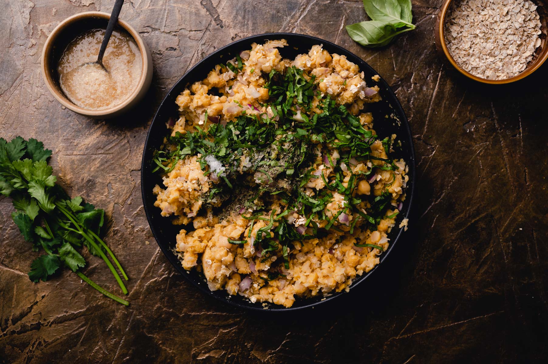 A skillet of risotto garnished with herbs and parmesan, alongside a bowl of sauce and a spoonful of oats, set on a dark, textured surface.