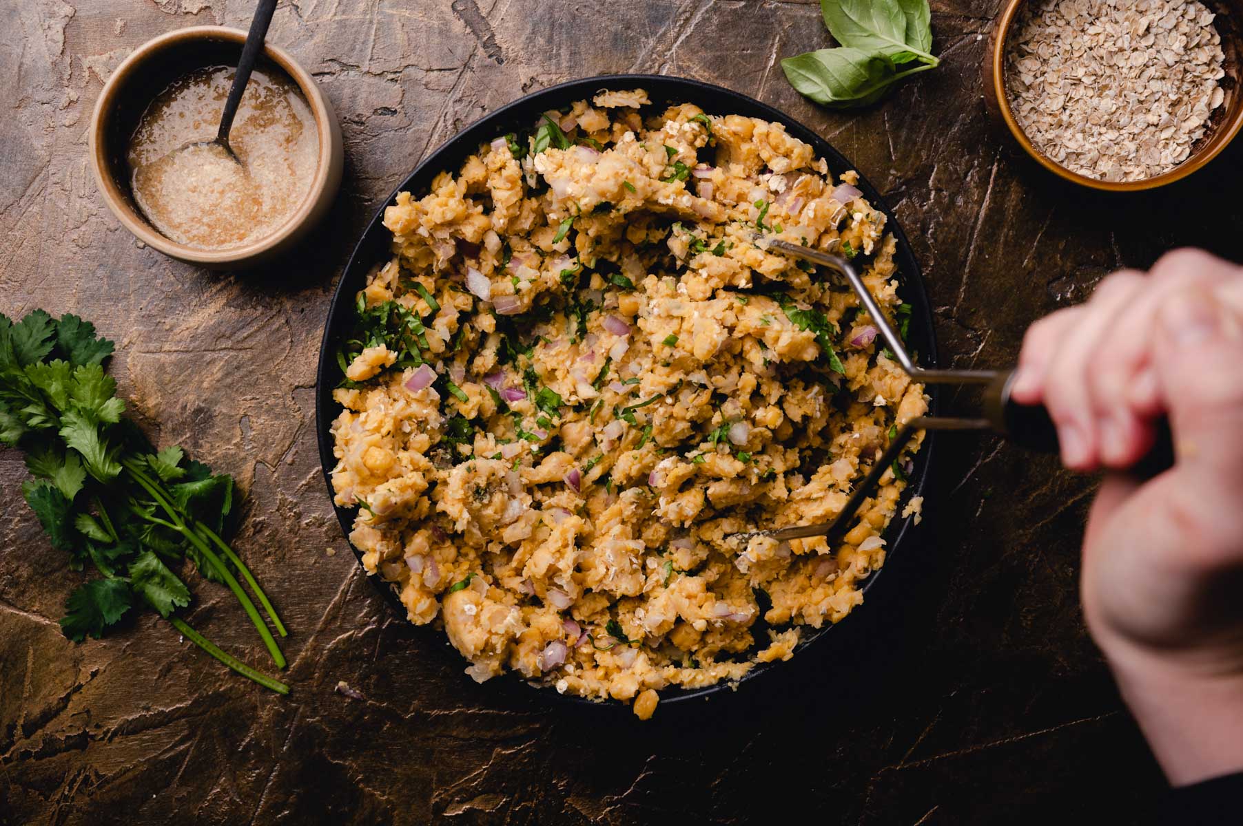 A person stirring a pan of cooked risotto garnished with herbs, with a bowl of sauce and oatmeal on the side.