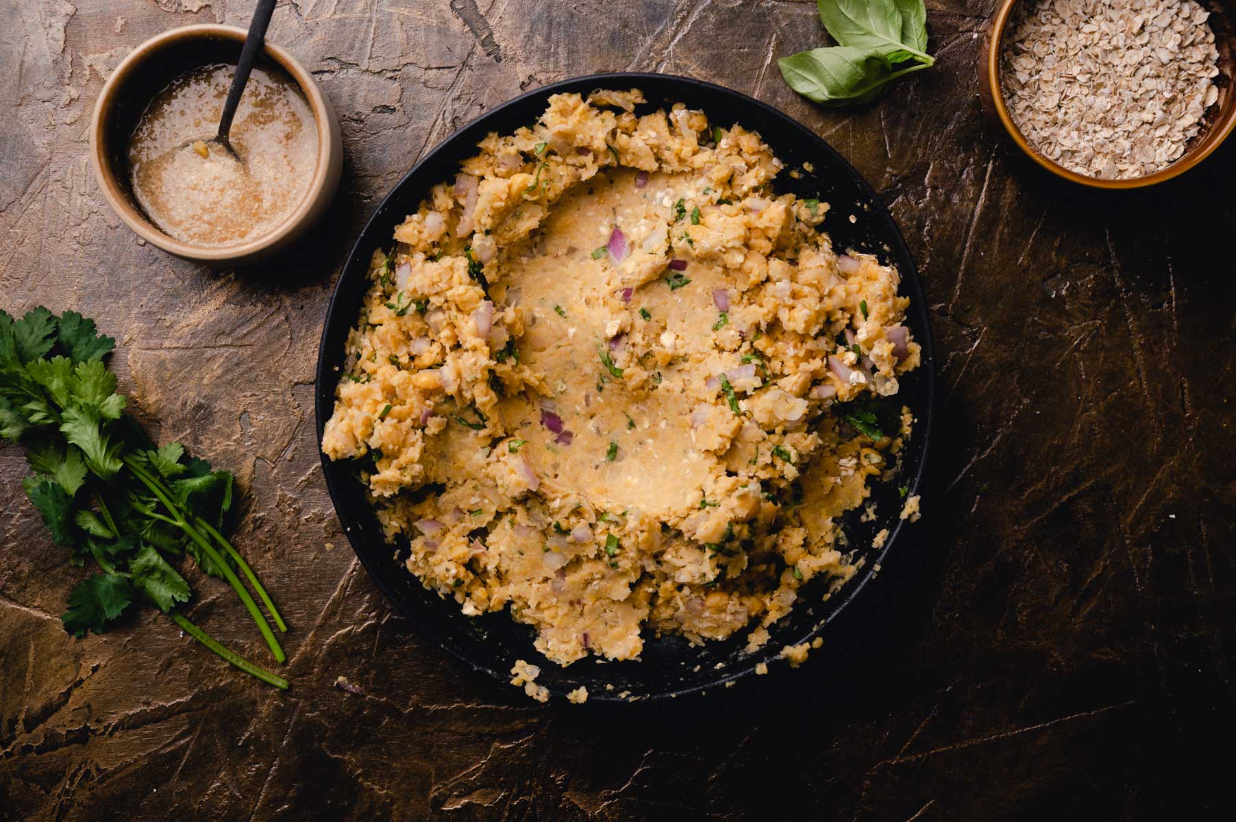 A skillet of creamy risotto garnished with herbs, accompanied by a sauce dish and fresh ingredients on a dark stone surface.