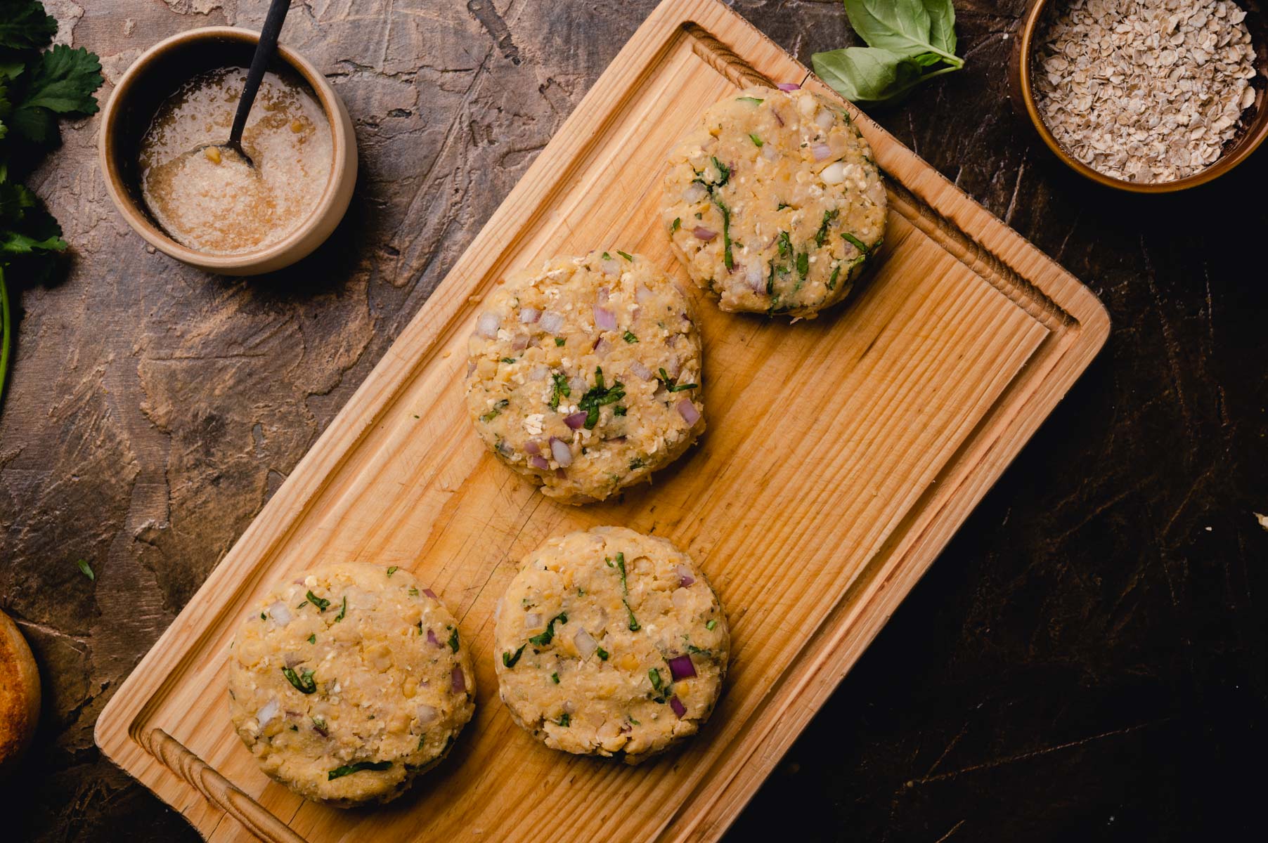 Four homemade veggie burgers on a wooden cutting board with a side of sauce and scattered ingredients like oats and parsley.
