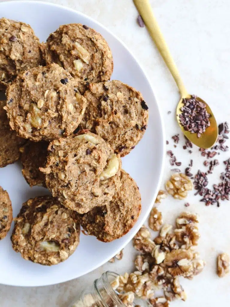 A plate of homemade oatmeal cookies with walnuts, accompanied by a spoonful of cacao nibs and scattered walnuts nearby, perfect for vegan muffin recipes.