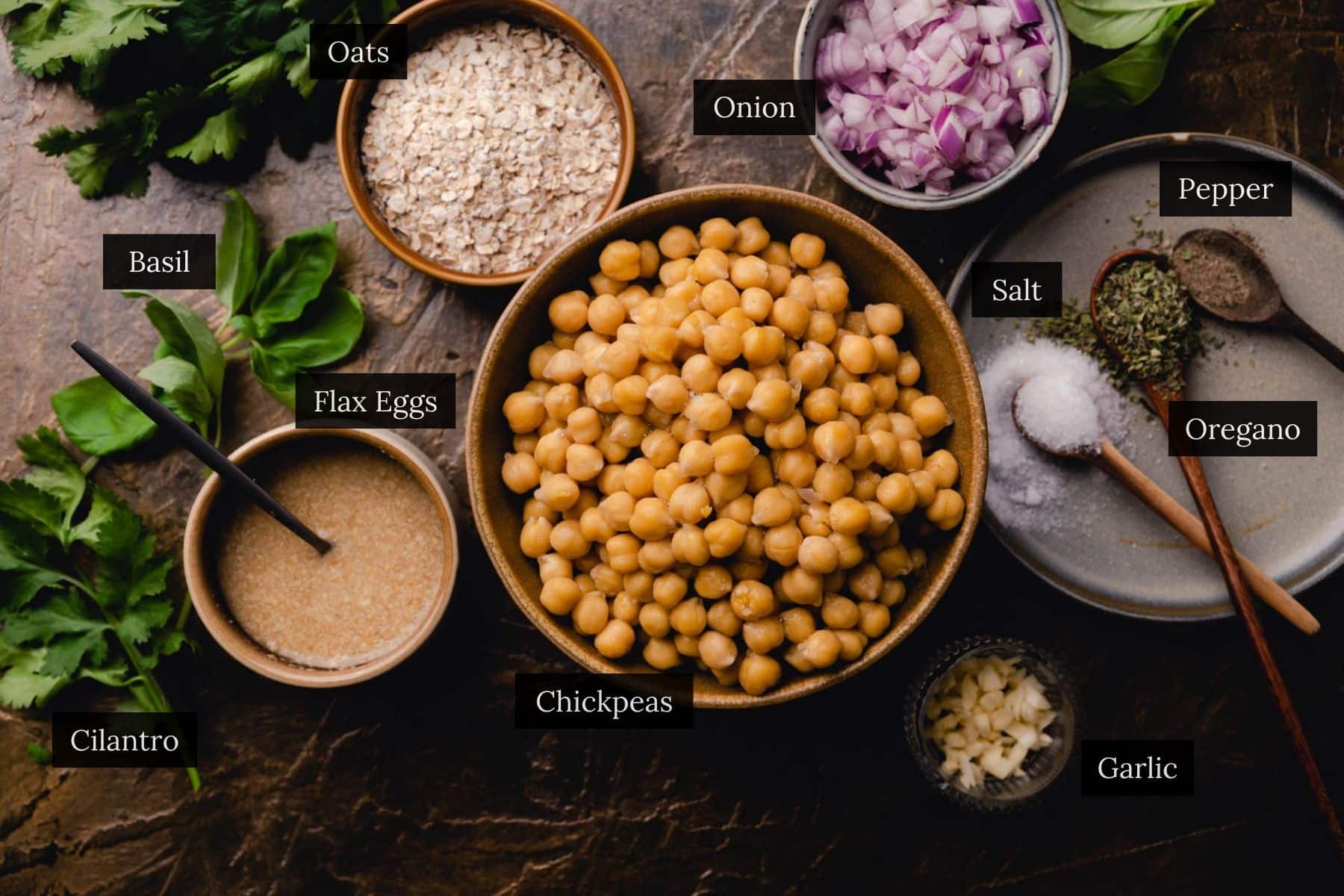 Top view of various ingredients placed on a dark surface, including chickpeas, oats, chopped onion, ground flax seeds, chopped garlic, salt, pepper, oregano, basil, and cilantro.