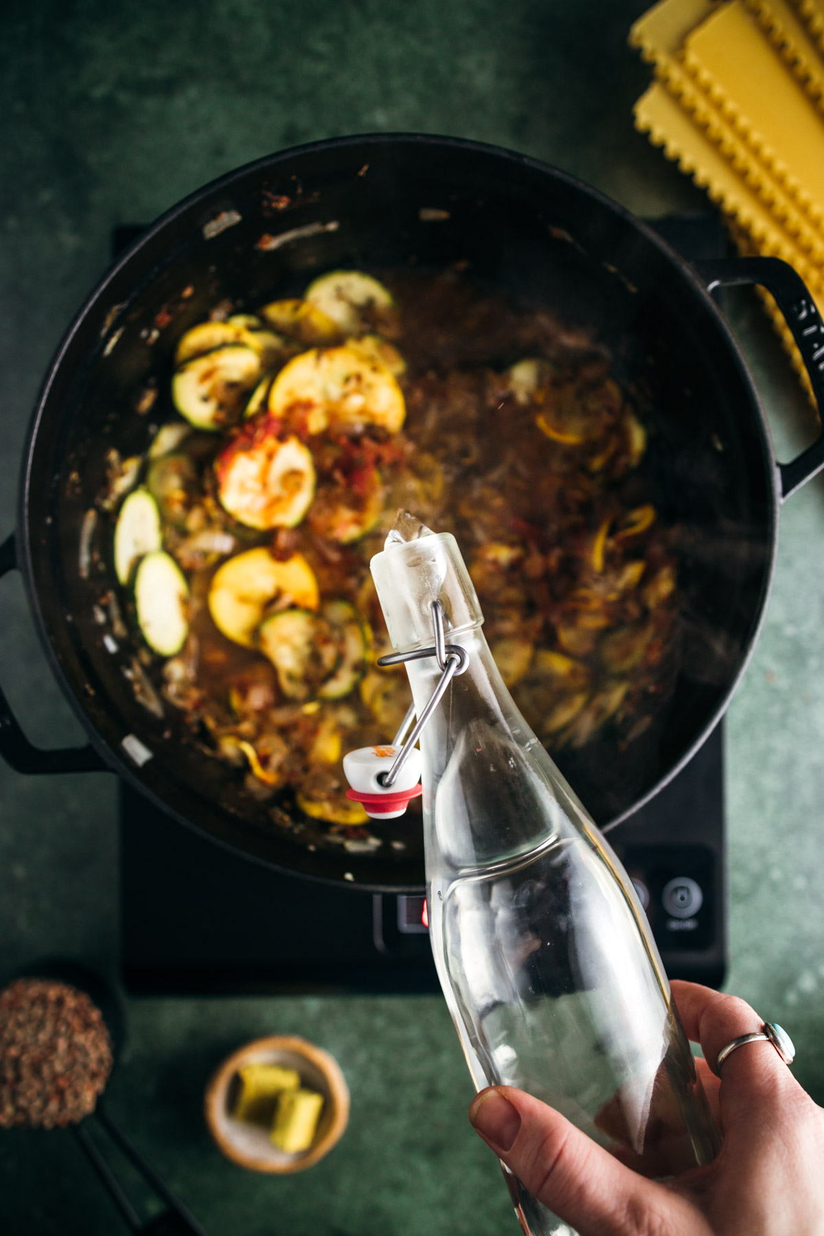 A person's hand holding a glass bottle with a clamp as they cook a dish with zucchini and tomatoes in a black pot on an induction stove.