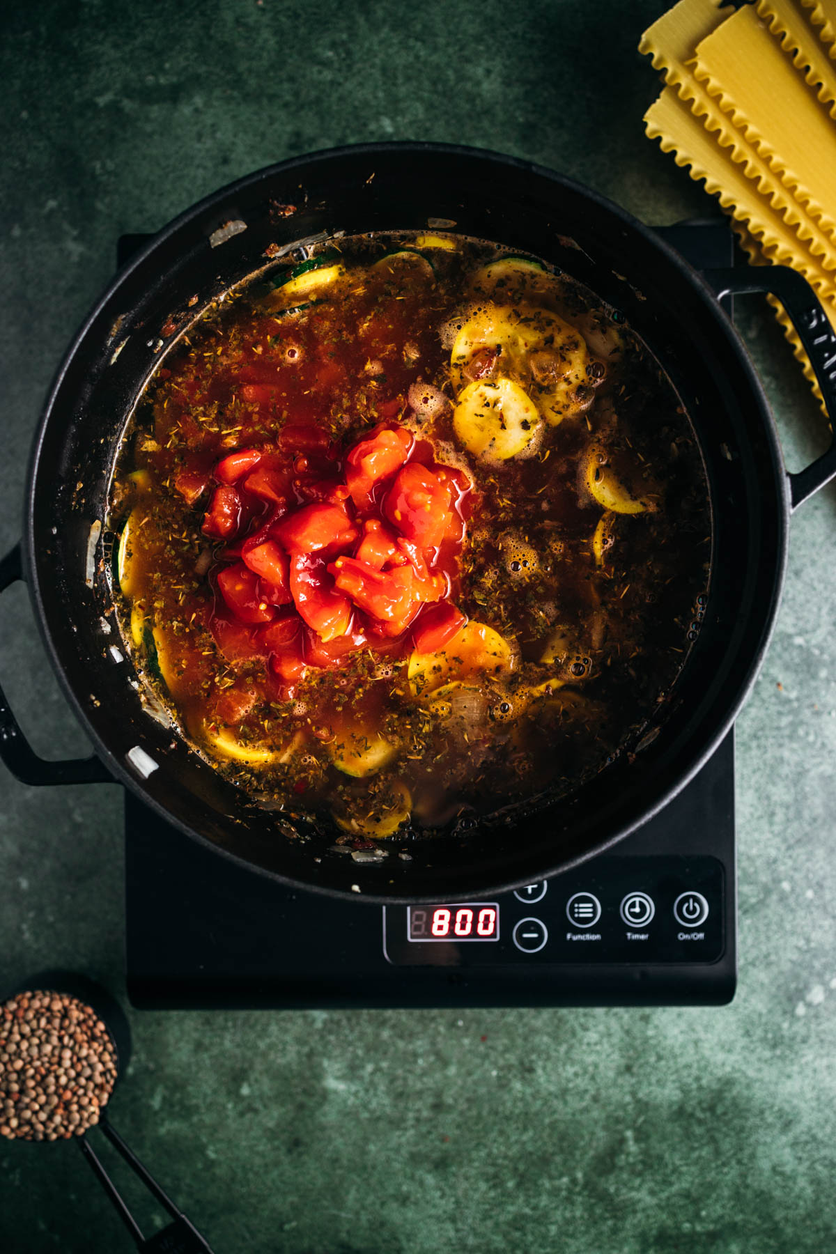 A slow cooker filled with soup containing tomatoes and herbs set on a digital display showing "8:00" with pasta and lentils nearby on a green surface.