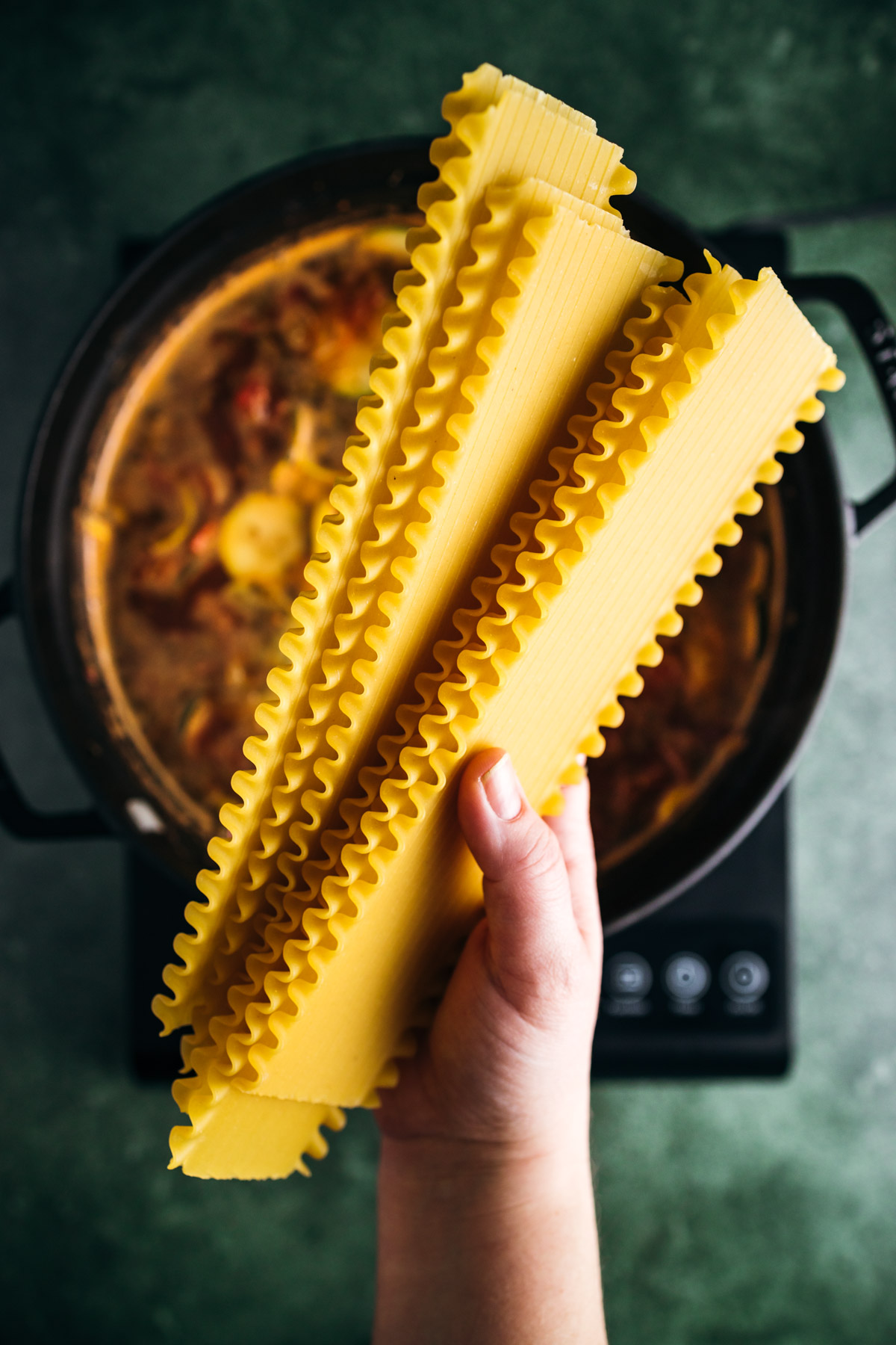 A hand holding uncooked lasagna noodles over a pot of simmering sauce on a stove.