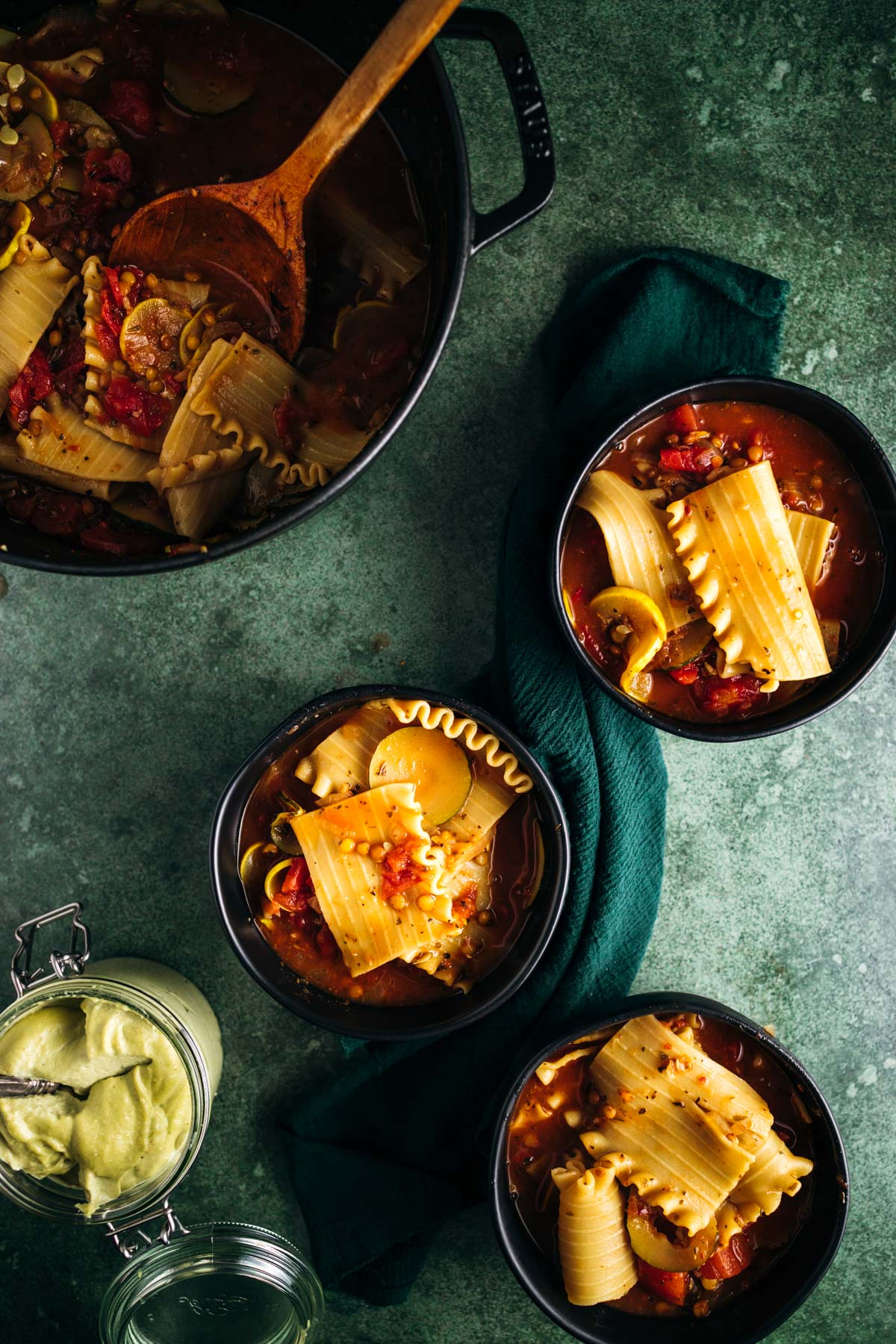 Three bowls of pasta soup with tomatoes and chickpeas, served on a dark textured surface, with a pot and wooden spoon alongside.