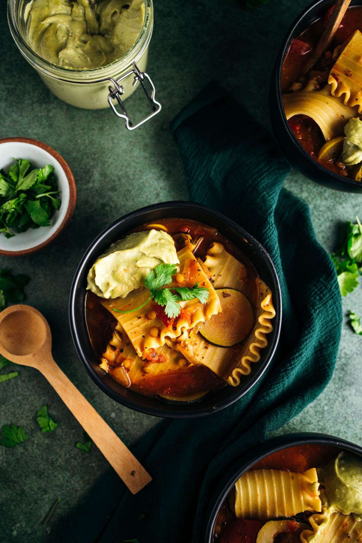 A bowl of pasta soup with a dollop of green sauce, garnished with a sprig of cilantro, alongside wooden utensils and fresh ingredients.