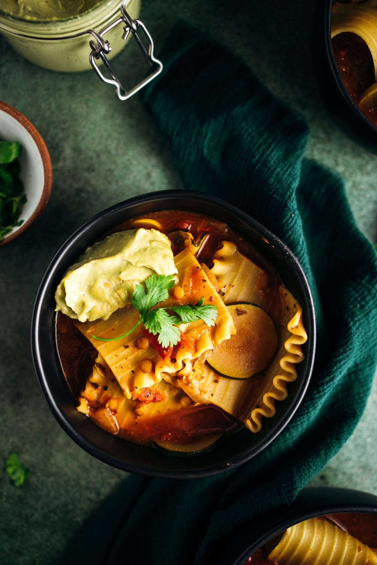 A bowl of lasagna soup garnished with a dollop of ricotta cheese and fresh cilantro, served on a dark green cloth.