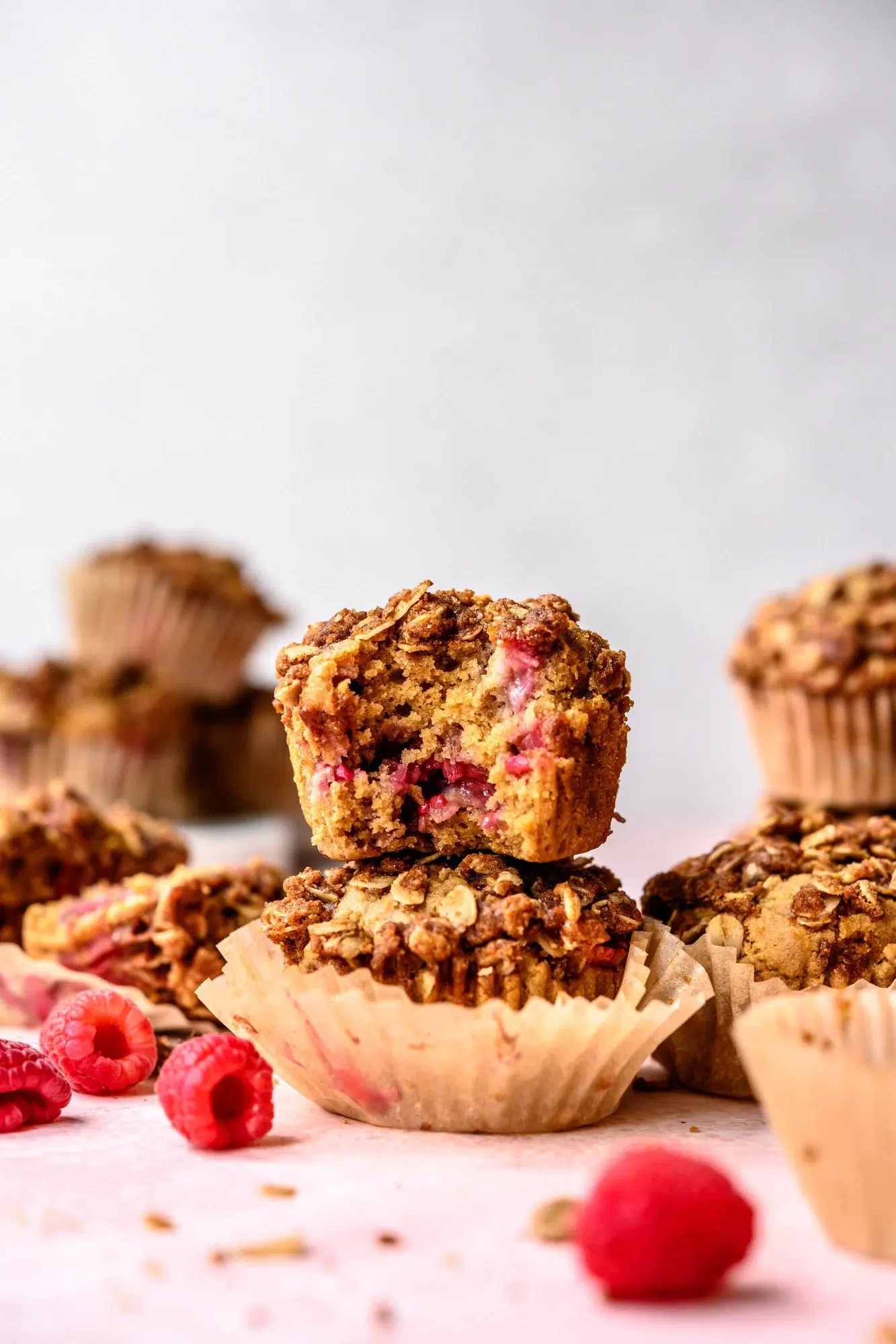 A vegan raspberry muffin with a bite taken out, surrounded by whole muffins and scattered raspberries, set against a light background.