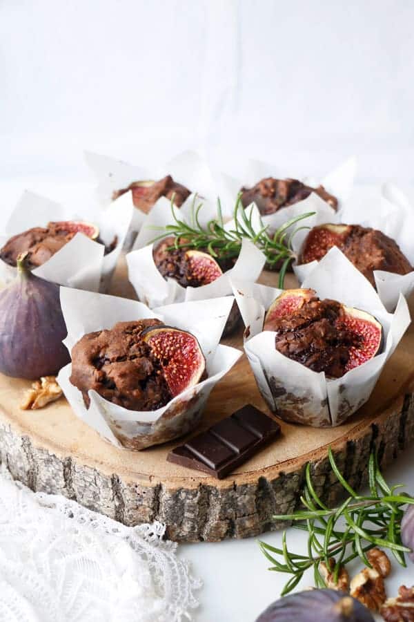 Vegan chocolate muffins topped with fig slices, presented on a rustic wooden board garnished with rosemary sprigs and walnuts, with pieces of chocolate on the side.