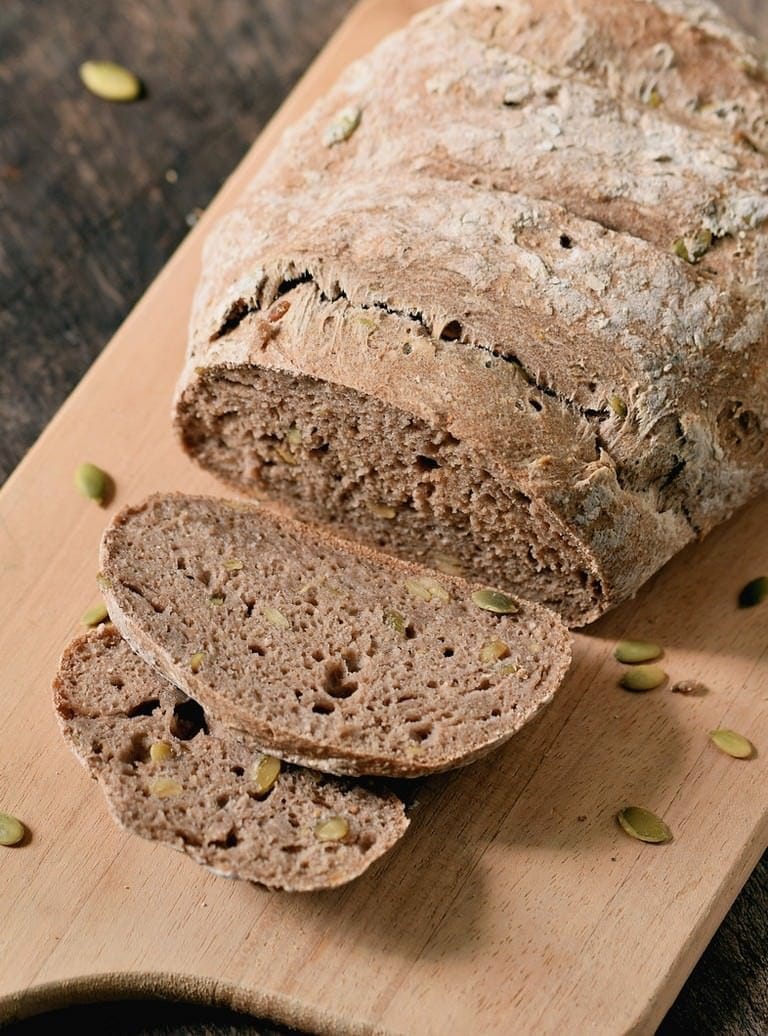 A loaf of multigrain bread with slices cut, on a wooden board scattered with seeds, perfect as a side dish.
