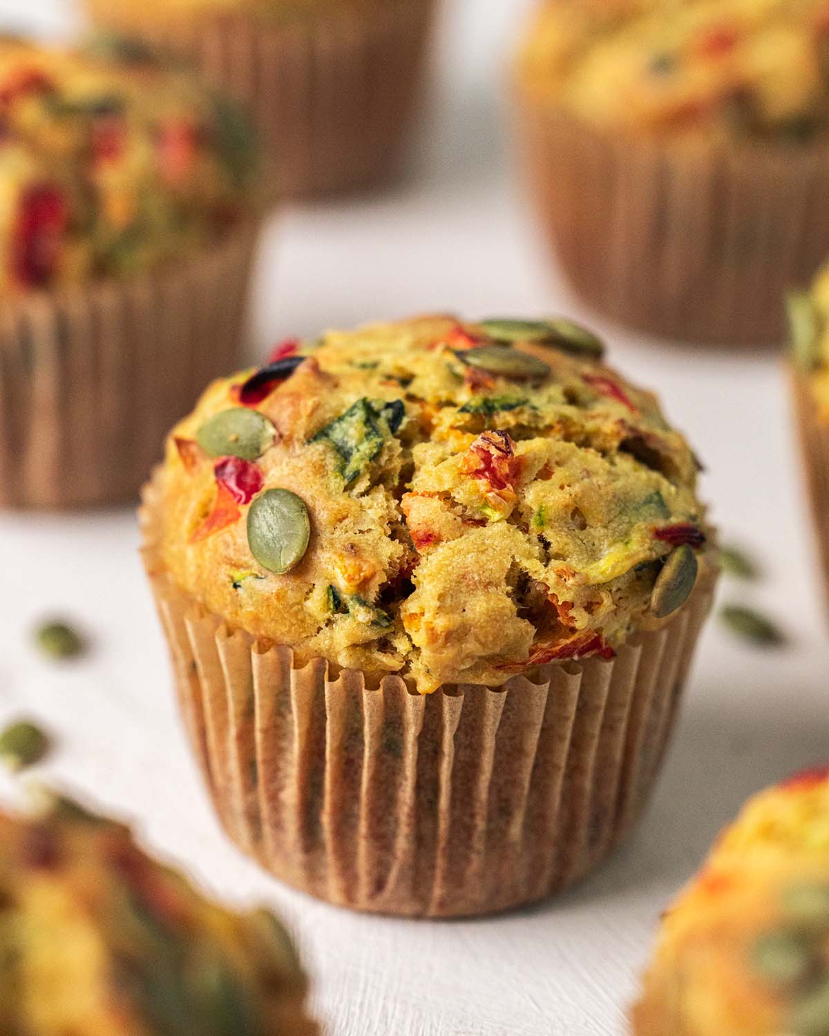 A colorful vegan vegetable muffin with visible chunks of red peppers and green onions, served in a brown paper cup on a light wooden surface.