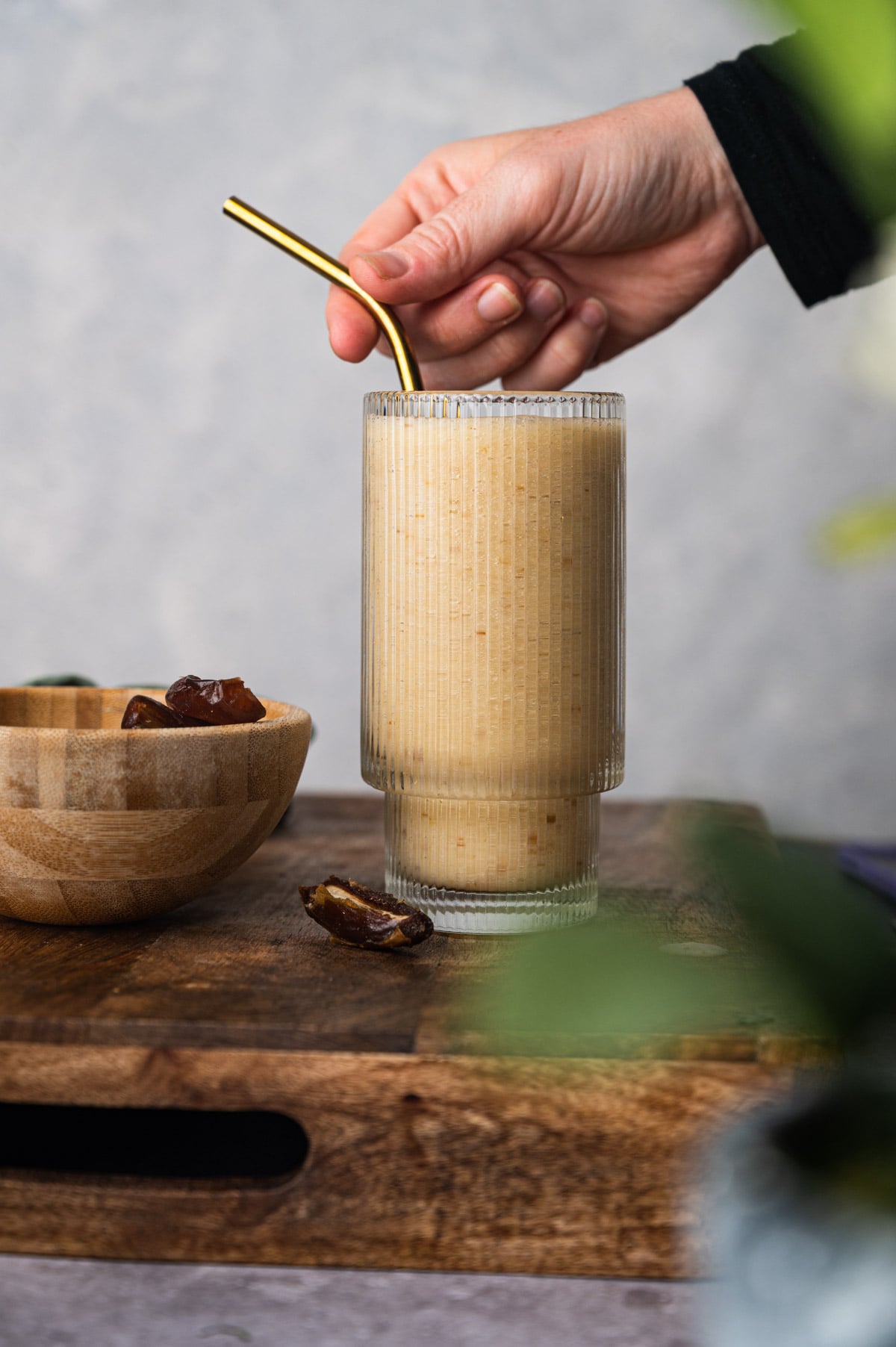 A hand holds a gold-colored straw in a glass of smoothie on a wooden surface, with a bowl of dates and a few scattered dates nearby.