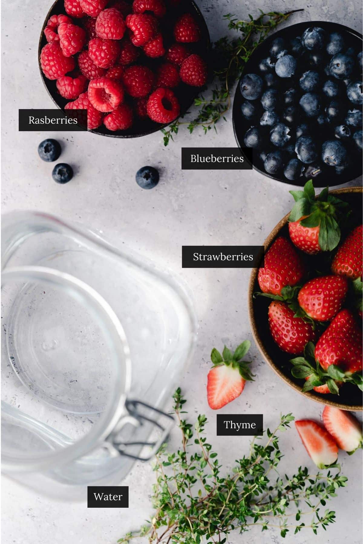 Bowls of raspberries, blueberries, thyme, and strawberries by a glass pitcher of water.
