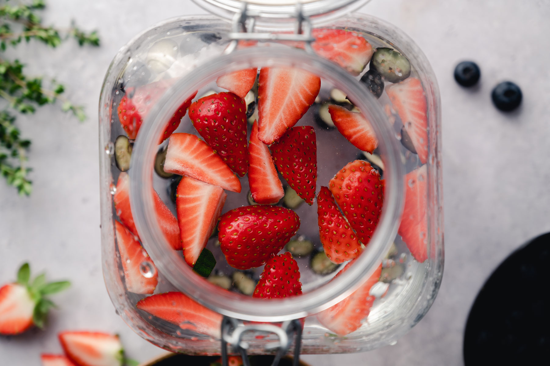 Glass jar filled with sliced strawberries and other fruits, viewed from above. A few blueberries and green herbs are visible around the jar.