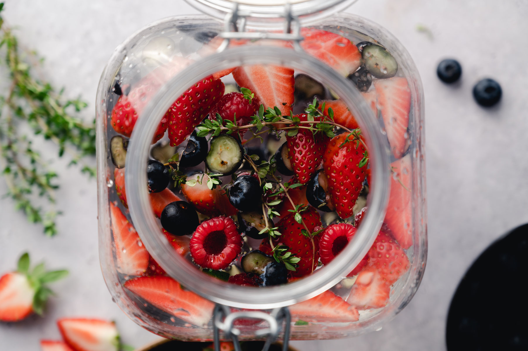 A glass jar filled with cut strawberries, blueberries, raspberries, and sprigs of thyme viewed from above. Some berries and thyme are scattered around the jar.