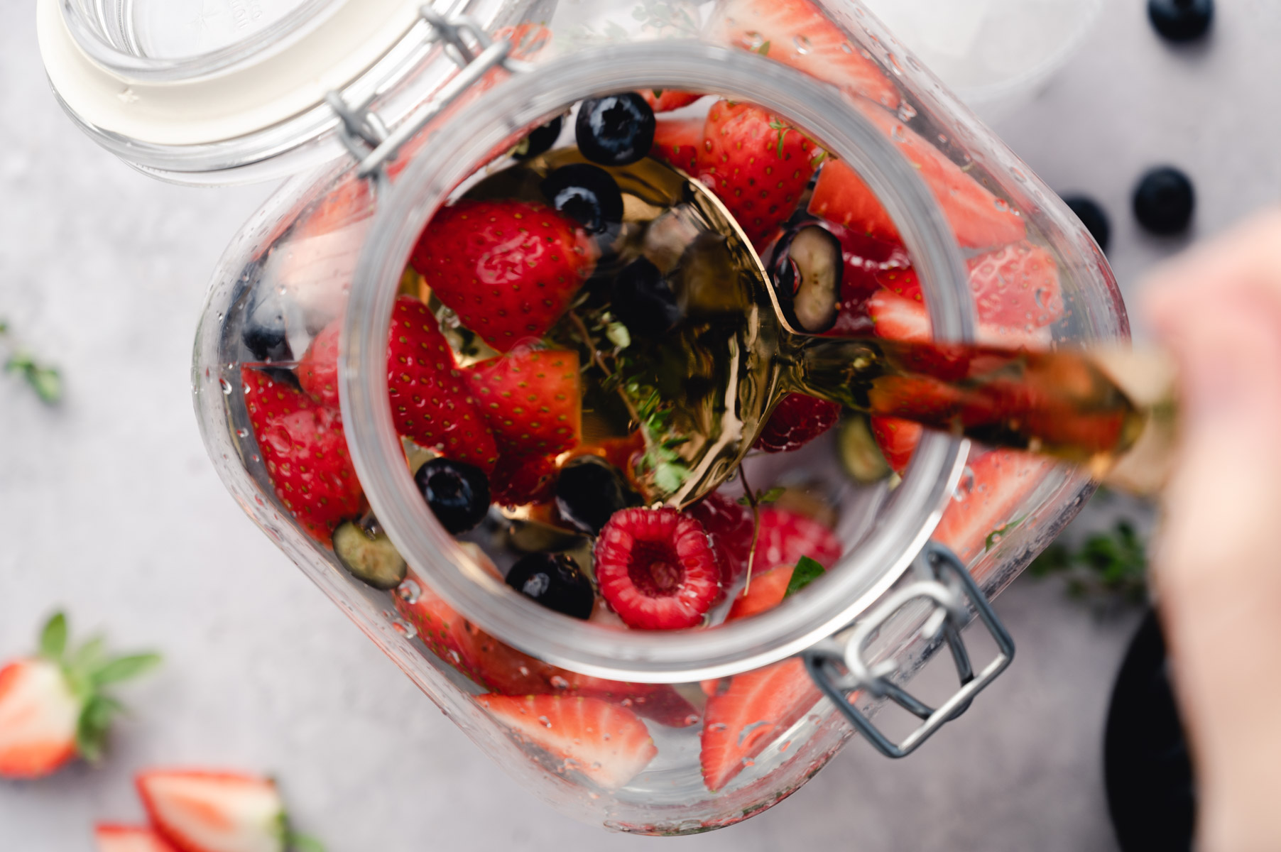Top view of a glass jar filled with sliced strawberries, raspberries, blueberries, and herbs, with a person stirring the contents with a wooden spoon.