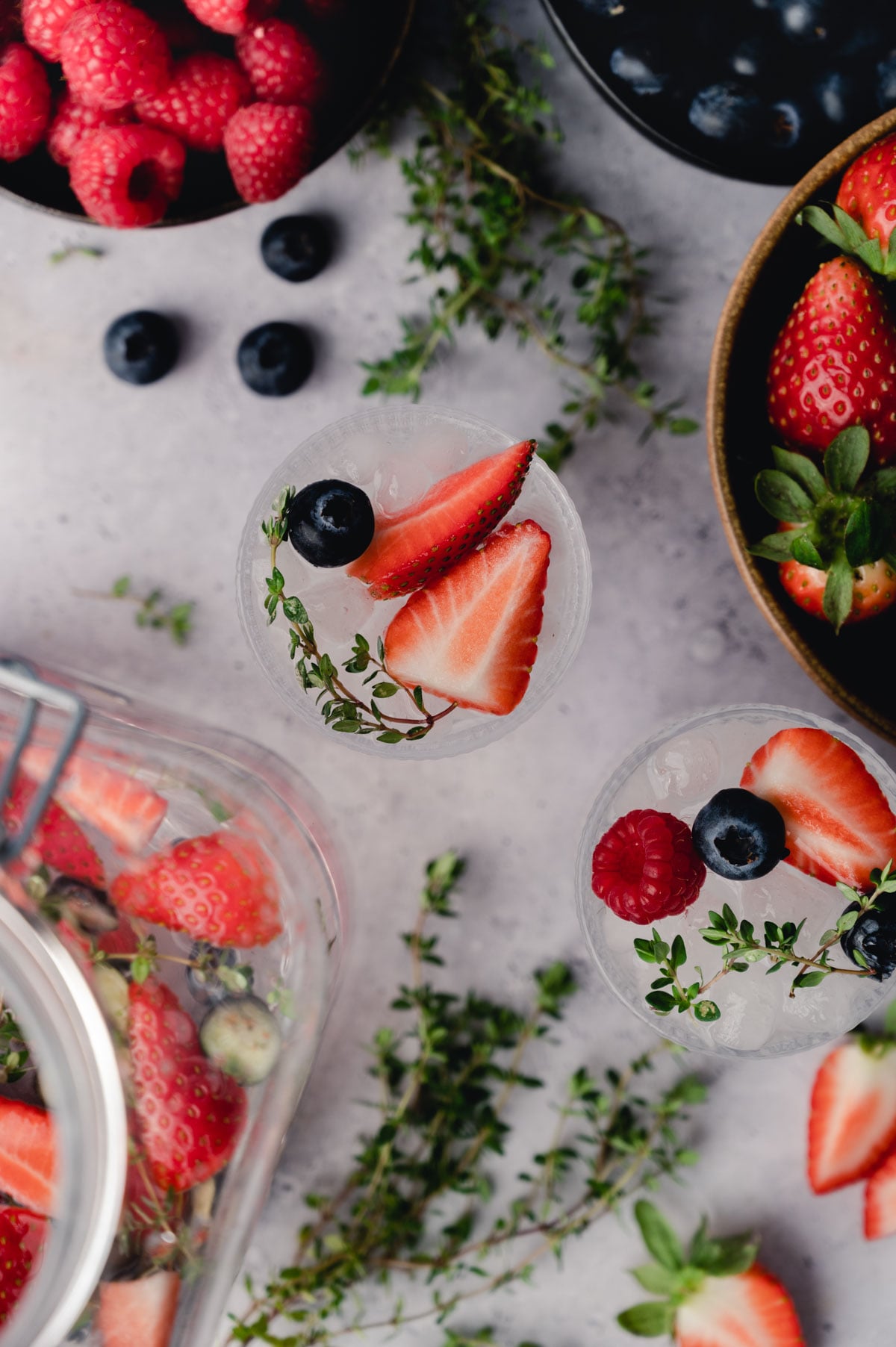 A top view of a table with various berries and herbs. Two clear glasses contain drinks garnished with strawberries, raspberries, blueberries, and sprigs of thyme.