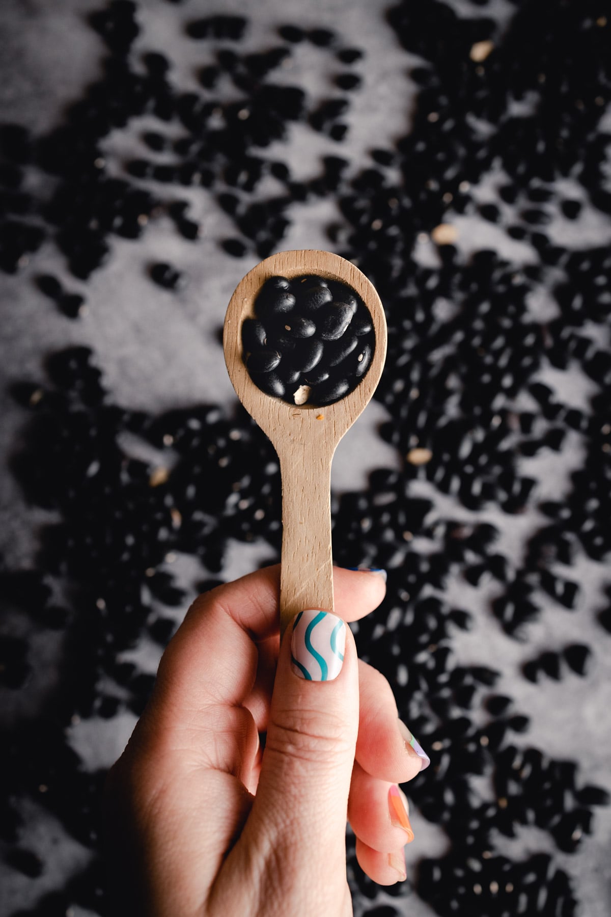 A hand with nail art holds a wooden spoon filled with black beans, with more black beans scattered in the background.