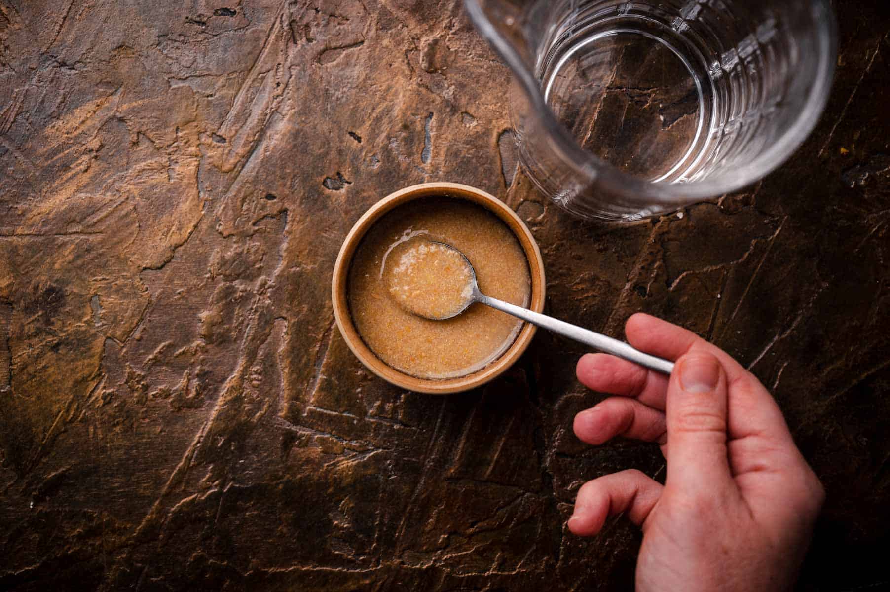 A hand holding a spoon stirs a brownish flax egg mixture in a small bowl next to a glass of water on a textured brown surface.