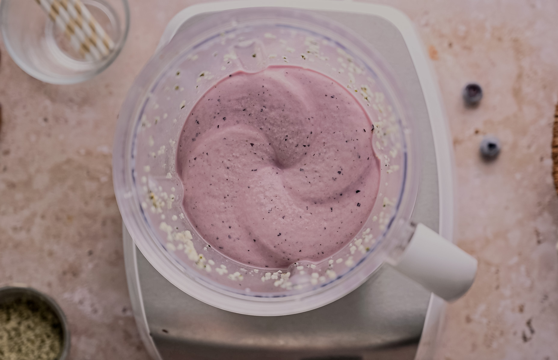 Top-down view of a blender with freshly made blueberry smoothie, surrounded by scattered blueberries and oats on a kitchen counter.