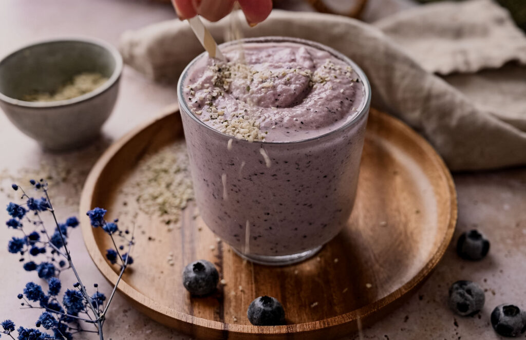 A hand holding a spoon over a glass of blueberry smoothie garnished with hemp seeds, on a wooden plate surrounded by blueberries and a bowl of seeds.