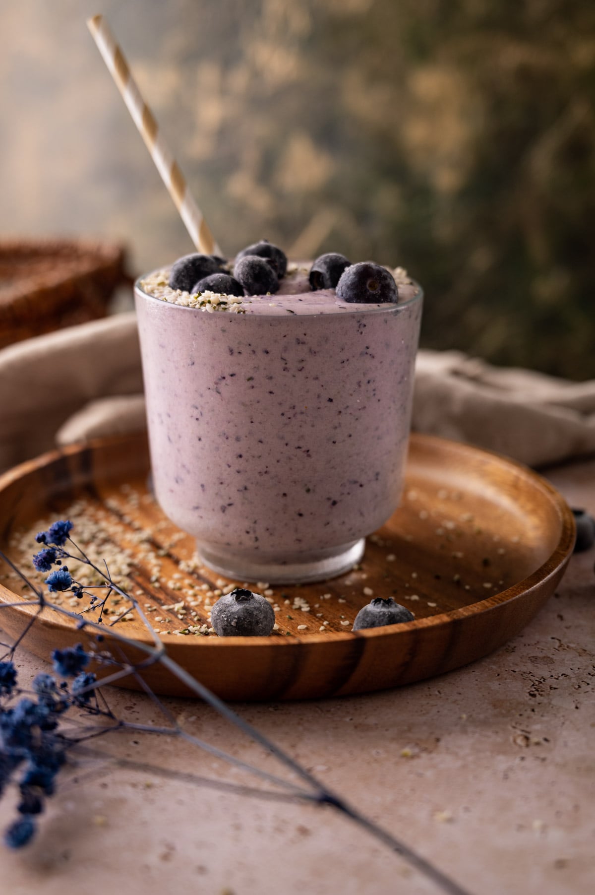 A blueberry smoothie in a clear glass, topped with whole blueberries and oats, served on a wooden tray with a slice of bread and decorative flowers.