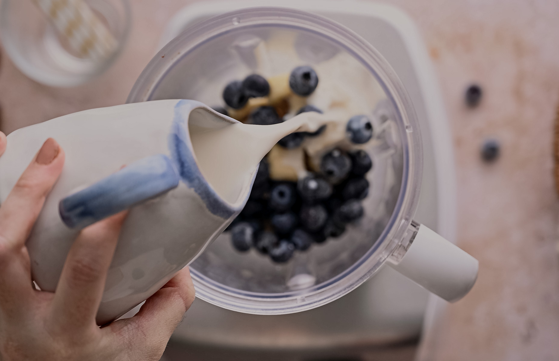 A person pours milk from a ceramic jug into a blender containing blueberries and bananas, with a marble countertop background.
