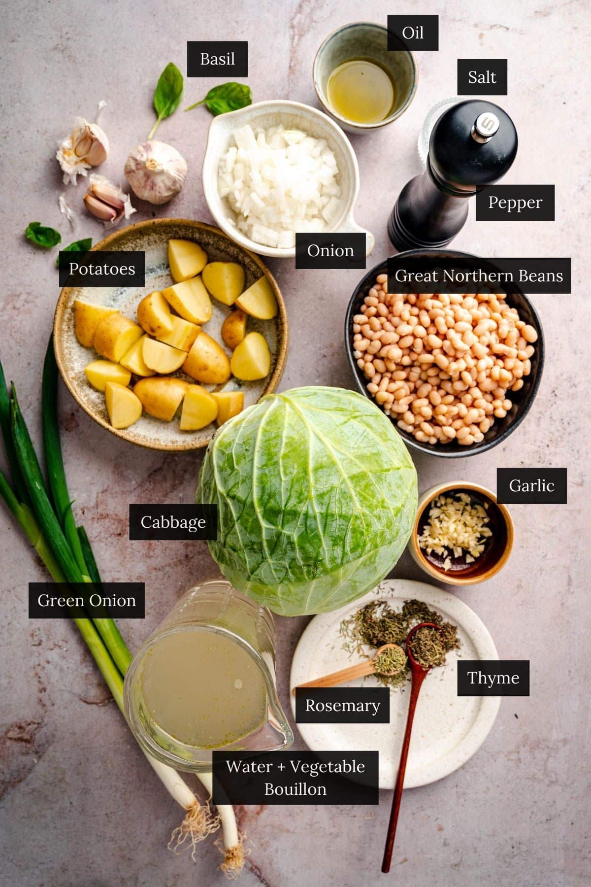 Assorted ingredients, perfect for a hearty cabbage soup, include potatoes, Great Northern beans, cabbage, green onion, basil, garlic, onion, salt, pepper, rosemary, thyme, oil, and vegetable bouillon on a light surface.